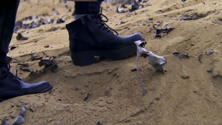 Close up of woman feet in black boots walking on sand with dry leaves. Female wearing leather black boots walking on sandy coast.の写真素材