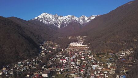Aerial view of a small town with mountain range with snowy top and blue clear sky on the background. Clip. Flying over the village located between mountains with forested slopes.の写真素材