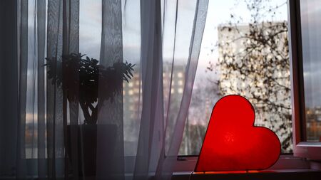 Bright lamp in shape of heart is on window. Concept. Red heart-shaped lamp stands at window and glows to bestow love on everyone. Bright romantic decor.の写真素材