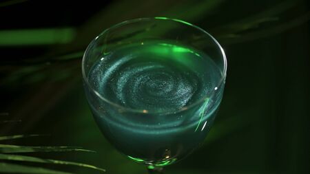 Wine glass with green coctail on dark green background with plant leaves. Stock footage. Close up of alcoholic beverage of green color with shining particles.の写真素材