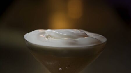 Putting cream to alcohol yellow cocktail with ice. Stock footage. Close up of adding to a cocktail in the martini glass a whipped cream on on dark blurred background.の写真素材