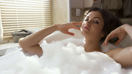 Young happy woman in the bath with white foam. Video. Portrait of a brunette female with curly hair...