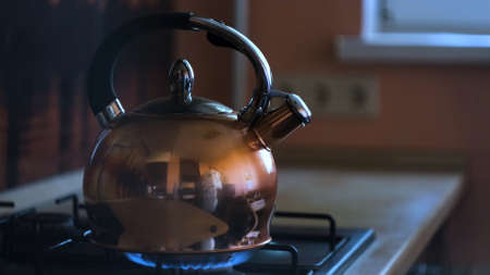 Close up of a stainless steel tea kettle on a flaming gas stove. Concept. Heating kettle with the reflection of the kitchen on its metal surface, preparation of hot beverage.の写真素材