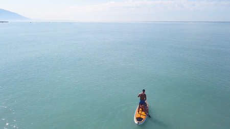 Aerial drone view of a man practicing stand up paddle or SUP in tropical exotic sea. Video. Flying over the turquoise calm sea and a man standing on a sup board on mountains and cloudy sky background.の写真素材