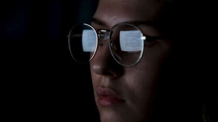 Beautiful girl works on internet with the reflection of a monitor inside glasses. Concept. Close up of woman face wearing glasses with the screen reflected in lenses isolated on black background.の写真素材