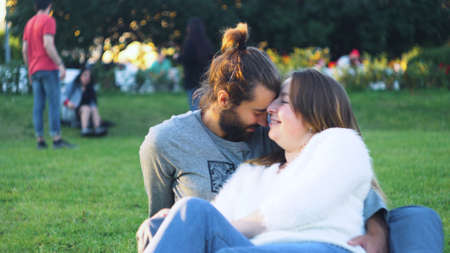 Beautiful couple caressing sitting on green grass in park. Concept. Happy young couple hugging on green grass in sun. Man and woman gently hug and smile sitting on grass in parkの写真素材