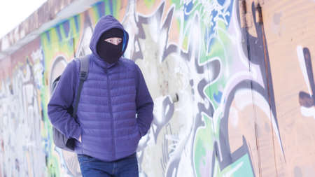 Serious young man in casual outfit and balaclava walking along the grafitti wall. Action. Male with a backpack hiding his face and looking around, concept of vandalism.の写真素材