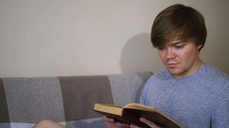 Young man is reading book intently sitting on sofa. Concept. Student reads classics with strained look. Man reads old book. Old classical literatureの写真素材