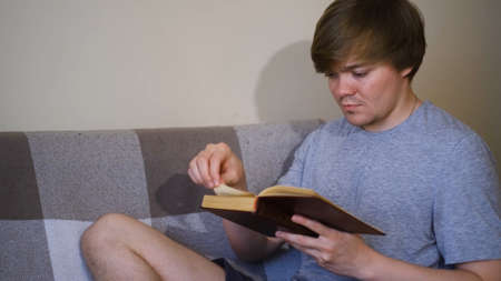 Young man is reading book intently sitting on sofa. Concept. Student reads classics with strained...