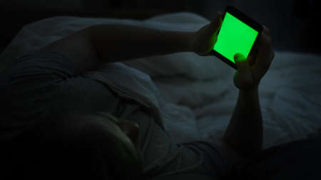 Man uses phone while lying in bed. Concept. Young man lying in bed uses smartphone with green screen. Dependence on smartphones and social networksの写真素材
