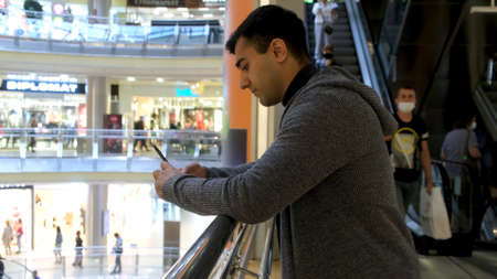 Side view of a young man standing in the shopping center and using his smartphone. Media. Male in casual clothes texting on the background of moving escalator.の写真素材