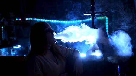 Female silhouette smoking hookah in bar. Media. Woman smokes hookah at bar in dark on background of...