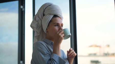 Woman with towel on head drinking coffee in background of window. Concept. Beautiful young woman...