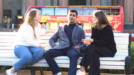 Three friends talking together while sitting on a white bench at the city park. Media. Two girls and a man smiling happily and enjoying time together.の写真素材