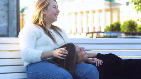Lesbian lying on the bench and the knees of her girlfriend in the park. Media. Lesbian woman kissing another girl on her forehead, laughing on a date, lgbt concept.の写真素材