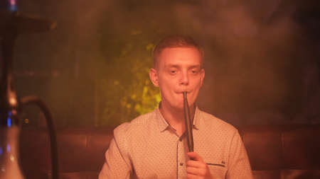 Man smokes a hookah on the dark background of a night bar or restaurant. Media. The pleasure of smoking on the face of a young man spending time while smoking shisha.の写真素材