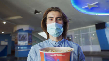 Young man in mask in cinema. Media. Handsome masked man holds popcorn outside movie theater. Going to cinema during pandemic. Coronavirus and movie theaterの写真素材