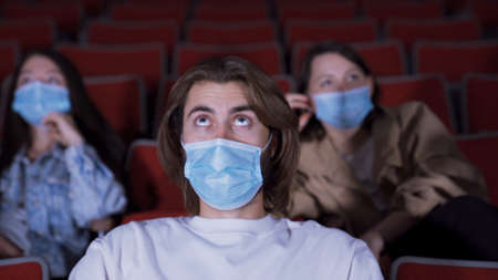 Man is tensely watching movie in movie theater. Media. Young man watches movie in fright while wearing medical mask. Going to movies during coronavirus pandemicの写真素材