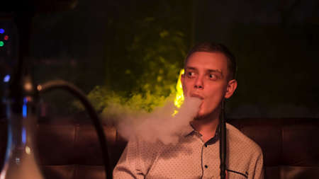 Fashionable modern young man smokes hookah in bar at night. Media. Blond guy holding tube of shisha, concept of smoking a hookah and having a good time.の写真素材