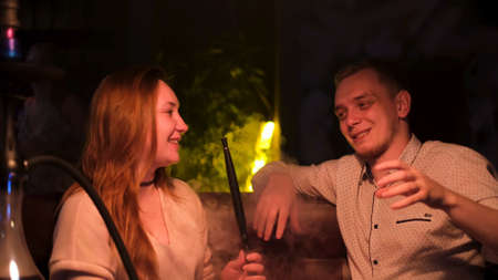 Couple smoking shisha in hookah bar. Media. Man and woman sitting on the couch, guy is making a joke to make her girlfriend laugh while she is holding a hookah tube.の写真素材