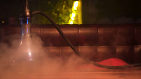 Glass hookah with glass bowl standing on the table in lounge bar. Media. Close up of shisha flask in clouds of smoke on a wooden table in a hookah cafe on the background of geen neon light lamp.の写真素材