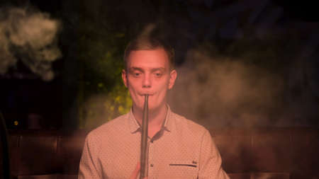 Young guy smokes hookah alone at the night bar and looking sad, concept of loneliness. Media. Blond male with bad habit, an alternative to tobacco.の写真素材