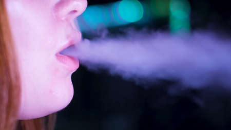 Close up of lips releasing vape smoke of e-cigarette or hookah on dark background. Media. Face details of a caucasian woman lips exhaling shisha smoke.の写真素材