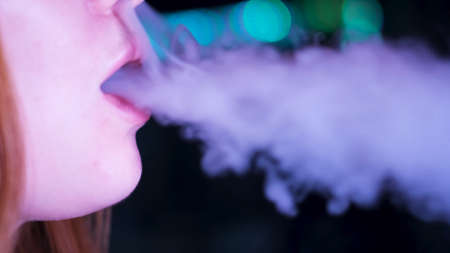 Close up of lips releasing vape smoke of e-cigarette or hookah on dark background. Media. Face details of a caucasian woman lips exhaling shisha smoke.の写真素材