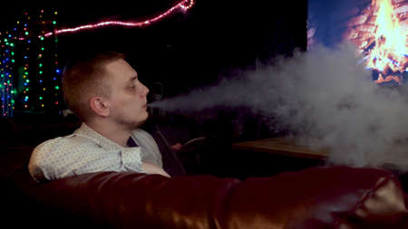 Side view of a man in relaxing atmosphere exhaling white thick smoke from his mouth. Media. Concept of nightlife and bad habit, male spending time at night bar.の写真素材