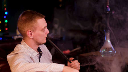 Side view of a man wearing white shirt smoking hookah and exhaling white smoke. Media. Young man...