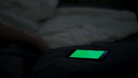 Close up of digital cell phone with green screen and a man in bed. Concept. Sleepy man using his smartphone and trying to sleep, concept of phone addiction and insomnia.の写真素材