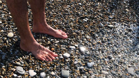 Barefoot male feet stands on a pebble beach washed by the sea. Concept. Close up of man legs and feet under water, pebbles visible through transparent waves.の写真素材