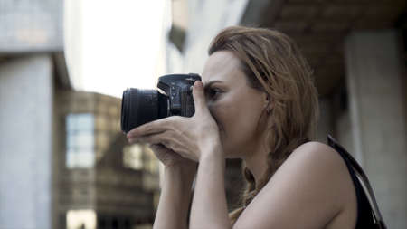 Female photographer with professional camera. Action. Young woman holding camera in her hands and taking pictures on street walk on glass facade building blurred background.の写真素材