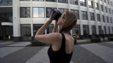 Beautiful young photographer woman takes photos outdoors of a modern business district. Action. Camera moving around a beautiful slim woman in black dress with a professional camera in her hands.の写真素材