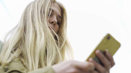Bottom view of young attractive woman using phone outdoors. Media. Female with long hair typing text...
