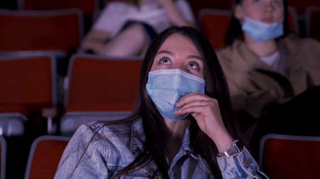 Portrait of a young beautiful girl with big green eyes watching movie in cinema with medical mask....