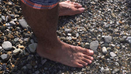 A sea waves on a stone beach covering the rocks and male feet. Media. Close up of man standing on the sea shore, summer rest and vacancy concept.の写真素材