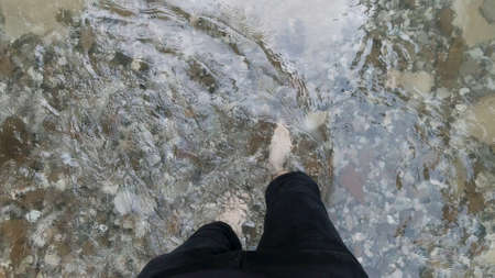 Man feet with hairy legs crossing cold mountain stream. Clip. Top view of male bare legs walking through cold river with stony bottom, concept of hiking.の写真素材