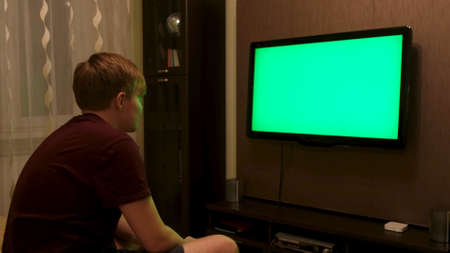 Man sitting on the couch in the living room and watching TV with green screen. Concept. Side view of young man turning off TV with chroma key and walking away.の写真素材