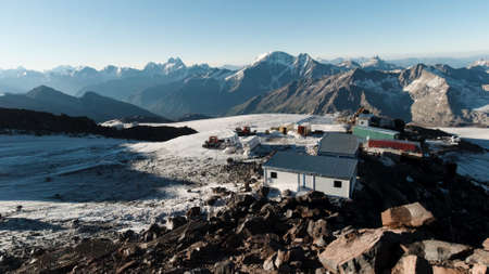 Natural landscape with snowy rocks and shelter for hikers. Clip. View from the hill top on a small camp base for climbers on the background of mountain range and blue clear sky background.の写真素材