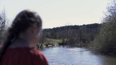 Rear view of a middle aged natural woman in a red shirt with long braid in front of flowing river...