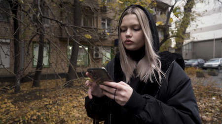 Portrait of a young blond girl typing on her smartphone device outdoors in autumn weather. Action. Beautiful teenager with a phone messaging in the street.の写真素材