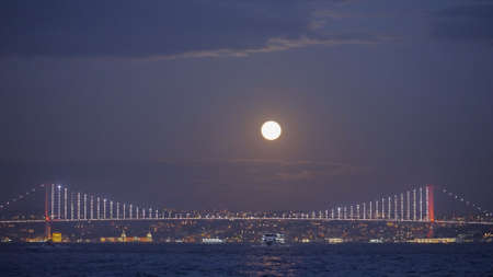 Sea landscape with shining city on background of evening sky with moon. Action. Beautiful coast on horizon with shining evening city. Moon over evening lights of city by seaの写真素材