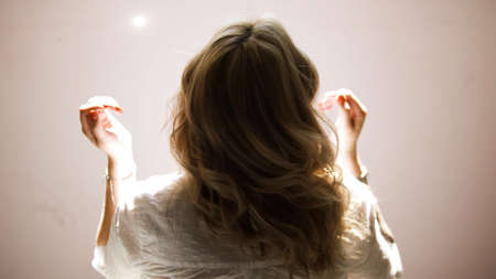 A young woman shaking her blond curly hair isolated on a beige wall background. Art. Rear view of a young blond woman in a white shirt posing and touching her hair.の写真素材