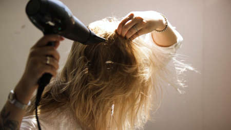 A young woman dries her hair with a hairdryer isolated on a beige wall background. Art. Young blond woman in a white shirt styling her hair.の写真素材