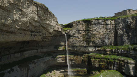 Narrow waterfall falls from high cliff. Action. Top view of water from hand falling into waterfall. River falls and becomes waterfall in mountains. Beautiful rocky cliff with narrow waterfallの写真素材