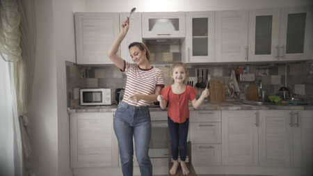 Happy family of two having fun in the kitchen together. Action. Playful young single mommy with overjoyed little preschool daughter holding forks and spoons and dancing.の写真素材