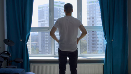 Young man admires view from window at home. Media. Rear view of a man with arms crossed looking through the window.の写真素材