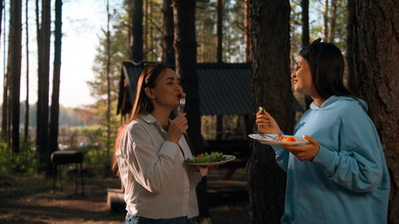 Barbecue on a summer day in coniferous forest. Stock footage. Two adult girls standing and communicating while eating.の写真素材