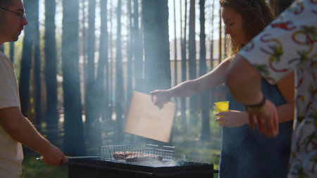 Friends cook meat on coals with grill. Stock footage. Woman blows up coals for grilling meat. Woman waves over grill in woods on sunny dayの写真素材